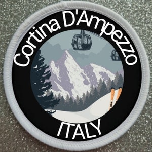 Peut inclure: Un écusson brodé circulaire à bordure blanche représentant un paysage de montagne, un télésiège et des skis orange. Le texte Cortina D'Ampezzo figure en haut et ITALY en bas en lettres blanches sur un cadre circulaire noir.