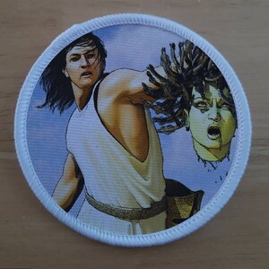 May include: A white circular patch featuring a colourful image of a man holding the severed head of a woman with snakes for hair. The man is wearing a white tunic and a gold belt. The woman's head is green and has a fierce expression.