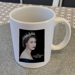 Könnte beinhalten: Eine weiße Keramiktasse mit einem Schwarzweißfoto von Königin Elisabeth II. mit einer Krone. Auf der Tasse steht der Text "RIP QUEEN ELIZABETH II 1926 - 2022".