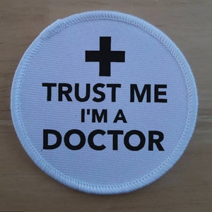 Puede incluir: Parche redondo de tela blanca con texto negro que dice "Trust me I'm a Doctor" y un signo más negro encima del texto.