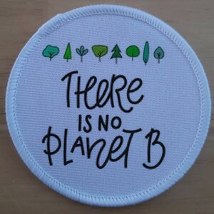 May include: White round patch with a black stitched border. The patch has a green and white illustration of trees at the top and the text "There is no Planet B" in black cursive writing.