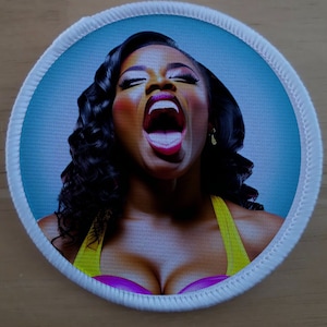 May include: A close-up of a woman with dark curly hair, wearing a yellow and pink top, with her mouth open and a surprised expression.