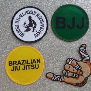 May include: Four embroidered patches: a white circular patch with black text reading "Jiu Jitsu It's All About Jesus and Acai" and a silhouette, a green circular patch with black "BJJ" text, a yellow circular patch with black "Brazilian Jiu Jitsu" text, and a tan hand gesture patch with white tape.
