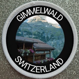 May include: A white and black circular patch with the text "Gimmelwald Switzerland" around the edge. The centre of the patch features a photo of a mountain hostel with a mountain range in the background.