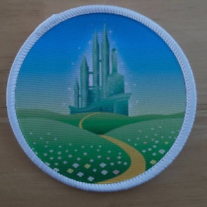 Puede incluir: Un parche redondo con borde blanco que muestra una imagen verde y amarilla de la Ciudad Esmeralda del Mago de Oz. La ciudad está en una colina con un camino de ladrillos amarillos que conduce a ella. El fondo es un cielo azul con nubes blancas.