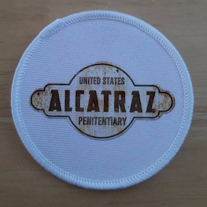 Puede incluir: Parche redondo de tela blanca con un gráfico marrón y amarillo de las palabras "United States Alcatraz Penitentiary".