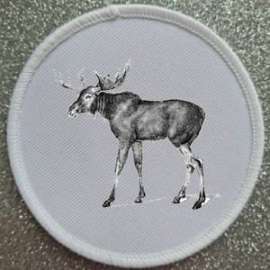 May include: A white round patch featuring a black and white illustration of a moose with large antlers. The moose is standing on a white background.