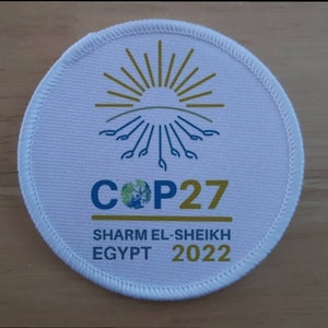 May include: White round patch with a gold sunburst design and the text "COP27" in blue and gold. Below the text is "SHARM EL-SHEIKH EGYPT 2022" in gold.