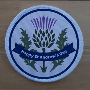 Könnte beinhalten: Ein runder weißer Aufnäher mit blauem Rand. Der Aufnäher zeigt eine grüne und violette Distel mit einem blauen Banner, auf dem "Happy St Andrew's Day" steht.