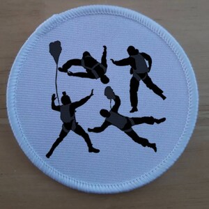 Könnte beinhalten: Runder weißer Patch mit einer schwarzen Silhouette von vier Fallschirmspringern in der Luft. Die Fallschirmspringer tragen Fallschirme und befinden sich in verschiedenen Positionen.