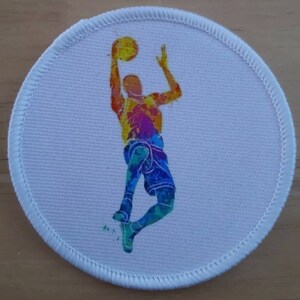 May include: A white round patch featuring a colourful silhouette of a basketball player making a jump shot. The player is wearing a jersey and shorts with a watercolour effect.