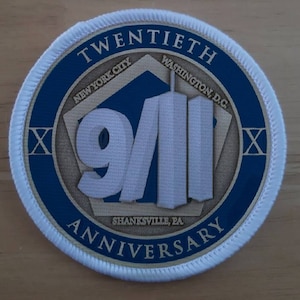 以下が含まれることがあります： 中央に青い円形がある白い丸いパッチ。円形には、白い数字「9/11」が書かれた金色の五角形があります。五角形には、「ニューヨーク市」、「ワシントンD.C.」、「シャンクスビル、PA」の文字が書かれています。青い円形には、「20周年記念」の文字が書かれています。