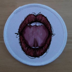 Puede incluir: Un parche redondo blanco con una boca abierta roja y rosa con una lengua que sobresale.