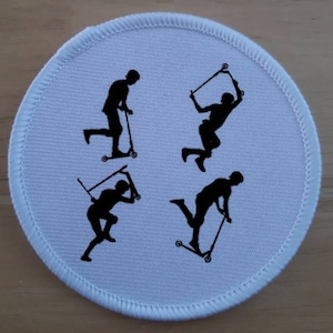 May include: White round patch with black silhouettes of four people riding scooters. The patch is made of a soft, textured fabric.