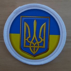 May include: A round, white patch with a blue and yellow Ukrainian flag design. The flag features a gold trident in the center.