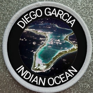 Pode incluir: Um remendo circular branco com uma borda preta e uma borda branca ao redor da borda externa. O centro do remendo apresenta uma imagem de satélite de uma ilha no Oceano Índico. O texto "DIEGO GARCIA INDIAN OCEAN" é impresso em branco ao redor da imagem.