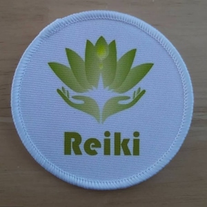 May include: White round fabric patch with a green lotus flower and two hands cupping the flower. The text "Reiki" is printed in green below the flower.
