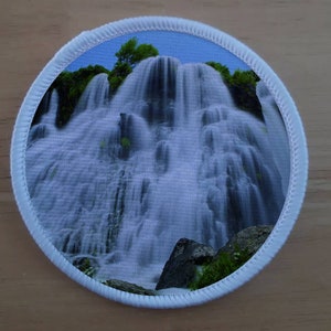 May include: A round white patch featuring a photograph of a waterfall cascading over rocks and green foliage. The waterfall is the main focus of the image, with the surrounding greenery and rocks providing context.