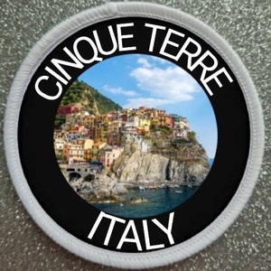 Peut inclure: Un écusson rond avec un centre noir représentant un village côtier coloré sous un ciel bleu. Les mots "CINQUE TERRE" et "ITALY" sont en blanc, courbés autour de l'image. L'écusson a une bordure blanche.