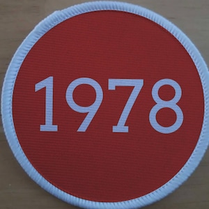 Può includere: Un cerotto circolare rosso con un bordo bianco e il numero 1978 stampato in bianco.