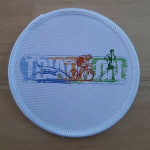Puede incluir: Parche redondo de tela blanca con un gráfico colorido de un triatlón. El gráfico muestra un nadador, un ciclista y un corredor con la palabra "TRIATHLON" en una fuente estilizada.