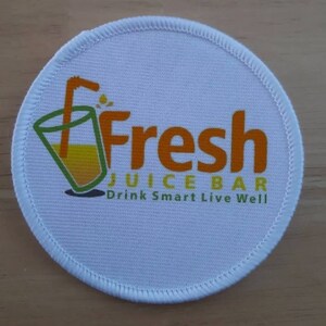 May include: White round fabric patch with a logo for a juice bar. The logo features a glass of orange juice with a straw and the word "Fresh" in orange letters. The text "Juice Bar" is in green letters below the word "Fresh". The text "Drink Smart Live Well" is in green letters below the words "Juice Bar".