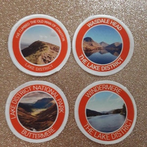 Könnte beinhalten: Vier runde Aufnäher mit Landschaftsbildern des Lake District. Jeder Aufnäher hat einen weißen Rand und einen orangefarbenen Ring mit Text. Die Aufnäher zeigen Bilder von Bergen und Seen, mit Text wie "Wasdale Head" und "Buttermere".