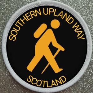 Op de afbeelding: Een ronde zwarte en witte patch met een witte rand. De patch heeft een geel silhouet van een wandelaar met een rugzak en een wandelstok. De tekst "SOUTHERN UPLAND WAY SCOTLAND" staat in het geel rond de wandelaar.