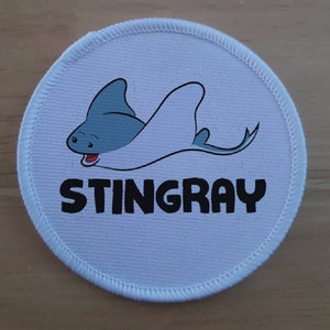 May include: A circular embroidered patch with a white background and a stitched border. The design features a cartoon blue and white stingray with a smiling face. Below the stingray, the word STINGRAY is printed in bold, black, capital letters.