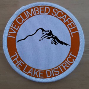 May include: White and orange round patch with the text "I've Climbed Scafell The Lake District" and a black silhouette of a mountain.