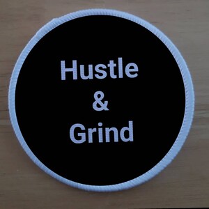Puede incluir: Un parche redondo negro y blanco con costuras blancas alrededor del borde. El texto "Hustle & Grind" está impreso en blanco sobre fondo negro.