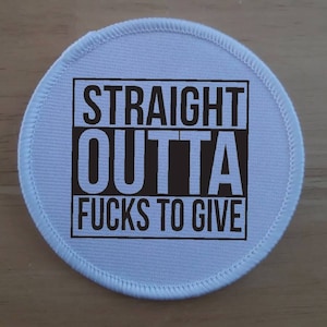 Peut inclure: Un écusson rond bleu clair avec le texte noir "STRAIGHT OUTTA FUCKS TO GIVE" dans un rectangle noir. Le patch est bordé d'une couture blanche.
