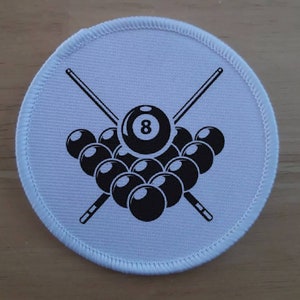 Puede incluir: Un parche blanco y circular con un diseño de billar en blanco y negro. El diseño incluye un conjunto de bolas de billar, la bola número 8 y dos tacos de billar cruzados. El parche tiene un borde cosido.