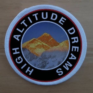 May include: A white, round patch with red and black trim. The patch features a mountain range with a sunrise in the background. The text "HIGH ALTITUDE DREAMS" is written in a circle around the image.