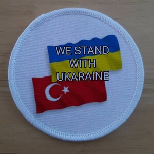 May include: A white round patch with the text "WE STAND WITH UKRAINE" and the flags of Ukraine and Turkey.