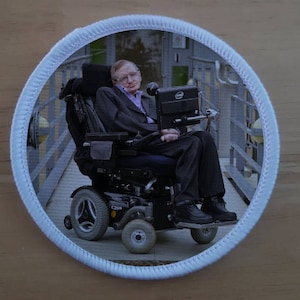 Puede incluir: Una foto en blanco y negro de un hombre en una silla de ruedas. Lleva un traje y una corbata. La silla de ruedas tiene un marco negro y ruedas. El hombre está mirando a la cámara.