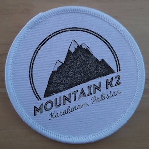 Op de afbeelding: Ronde witte patch met een zwart-witte afbeelding van een bergketen. De tekst "MOUNTAIN K2 Karakorum. Pakistan" is onder de afbeelding gedrukt.
