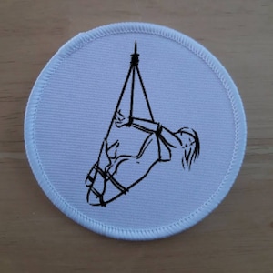 May include: A white round patch with a black line drawing of a person tied up and hanging from the ceiling.