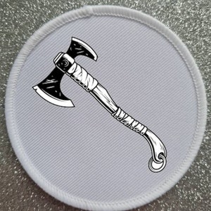 May include: A white round patch with a black and white illustration of an axe with a wrapped handle. The axe has a double-headed blade.