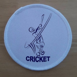 May include: White round fabric patch with a blue outline and a blue illustration of a cricket player batting. The text "CRICKET" is printed in blue below the illustration.