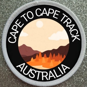 Può includere: Un cerotto circolare bianco con testo nero e un cerchio nero al centro. Il testo recita "Cape to Cape Track Australia". Il cerchio nero contiene un'illustrazione stilizzata di un paesaggio con montagne, alberi e un fiume.
