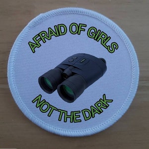 Peut inclure: Un écusson rond blanc avec du texte vert qui dit "Afraid of girls not the dark". Une paire de jumelles noires est au centre de l'écusson.