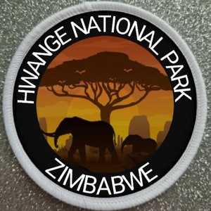 Könnte beinhalten: Ein weißer runder Aufnäher mit schwarzem Text und einem schwarzen und orangefarbenen Bild. Der Text lautet "Hwange National Park Zimbabwe". Das Bild zeigt eine Silhouette eines Elefanten, eines Elefantenbabys und eines Baumes vor einem Sonnenuntergang.