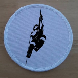 May include: A white round patch with a black silhouette of a person abseiling down a rope.