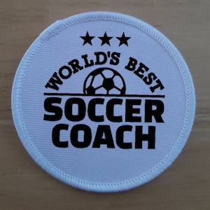 Może przedstawiać: Okrągła naszywka z białego materiału z czarnym tekstem "World's Best Soccer Coach" z trzema gwiazdami nad tekstem i piłką nożną w środku.