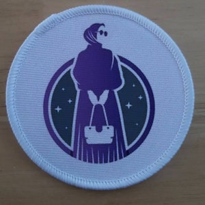 May include: A white round patch with a purple silhouette of a person wearing a headscarf and sunglasses, holding a handbag. The person is standing in front of a dark circle with white stars.