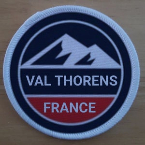 Könnte beinhalten: Ein runder weißer Aufnäher mit einem blauen und roten Kreis in der Mitte. Der Kreis zeigt eine weiße Bergkette und den Text "VAL THORENS FRANKREICH".