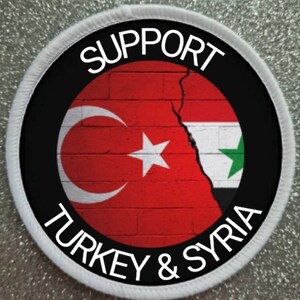 May include: A white circular patch with black text that reads "SUPPORT TURKEY & SYRIA". Inside the circle is a brick wall with a crack down the middle. The left side of the wall is painted with the Turkish flag and the right side is painted with the Syrian flag.