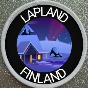 Puede incluir: Un parche circular blanco con texto negro que dice "Lapland Finlandia" alrededor de un círculo negro con una imagen colorida de una cabaña nevada con una aurora boreal púrpura en el fondo.