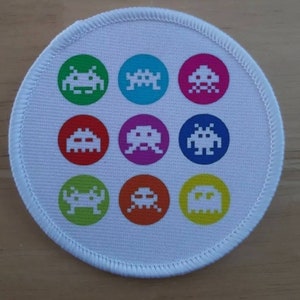 May include: A white round patch with nine colorful circles, each containing a pixelated alien design. The aliens are in different colors, including red, blue, green, yellow, and purple.
